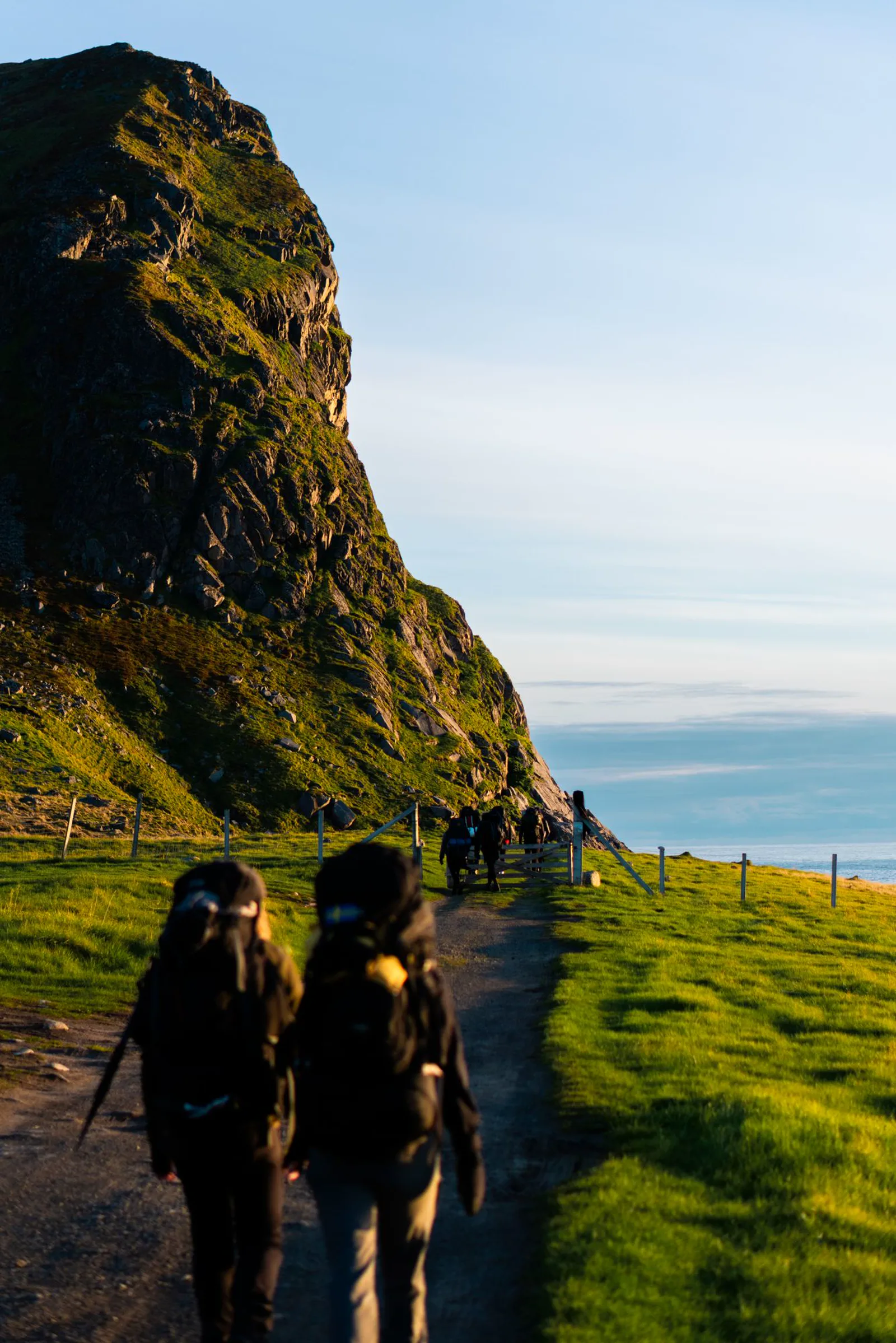Hikers on a coastal path beneath a steep green ridge at sunset